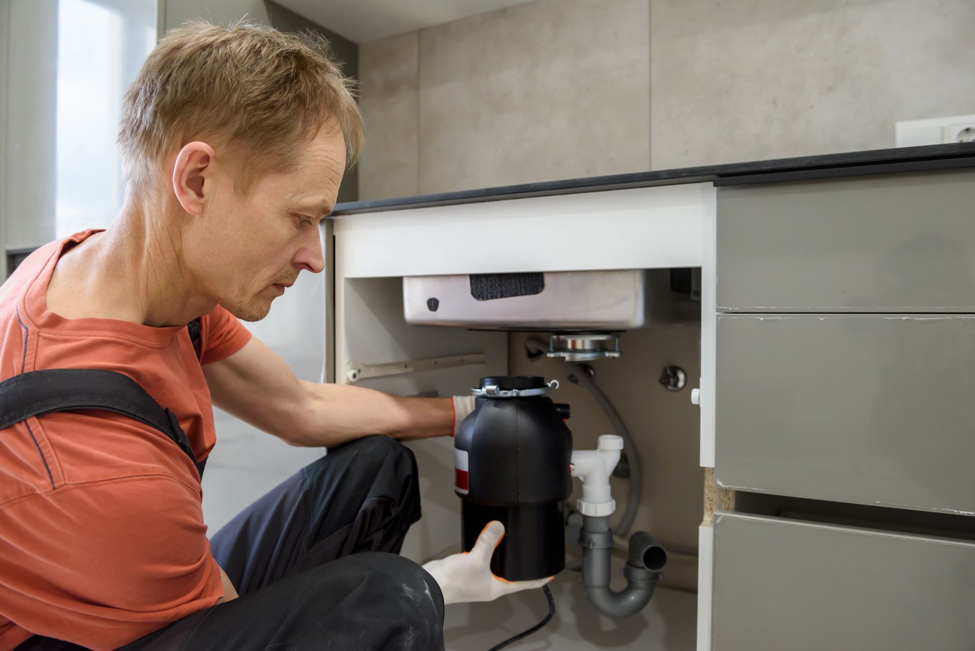 A man is fixing a garbage disposal under a sink in a kitchen.