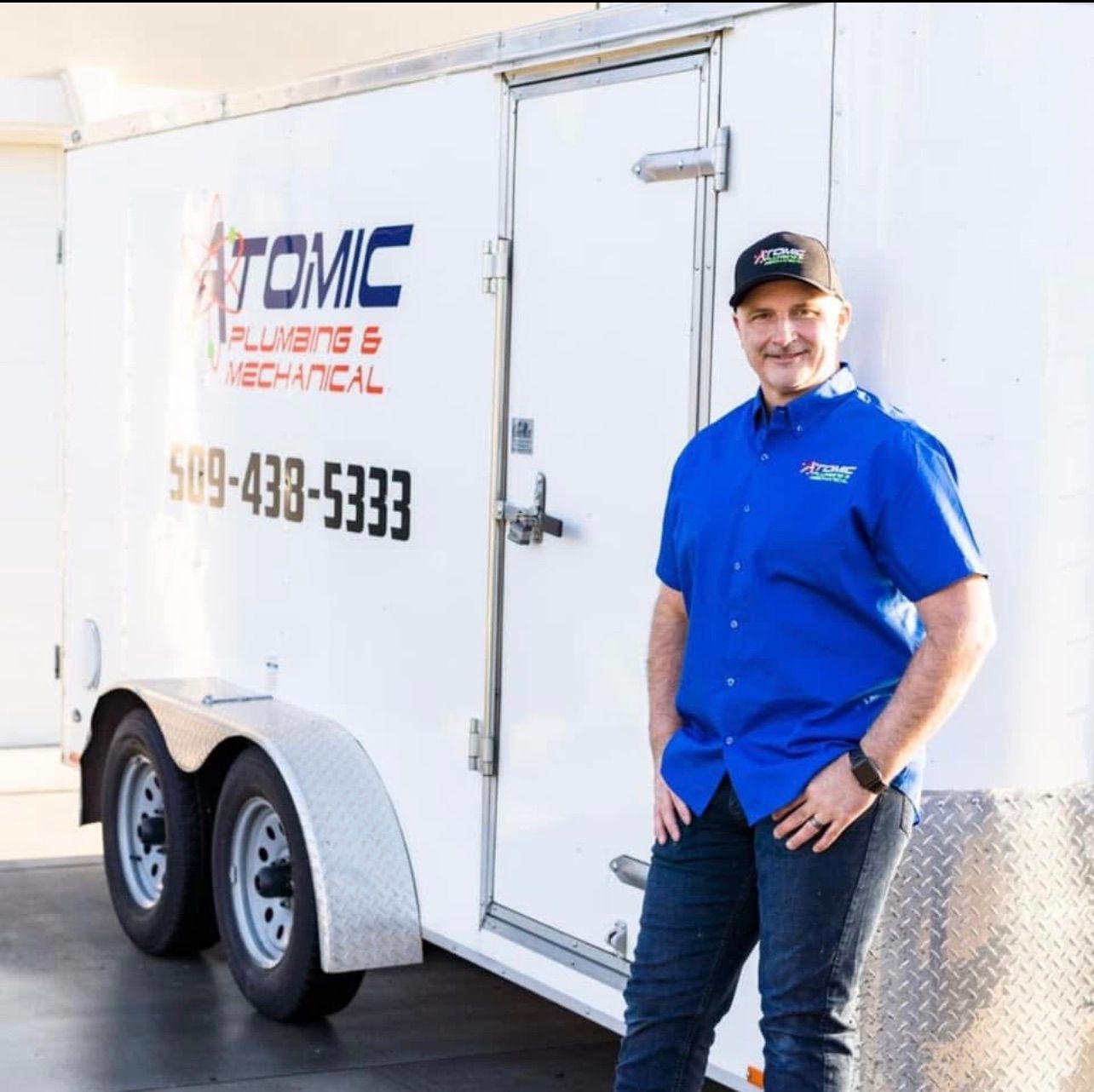 A man in a blue shirt is standing in front of an atomic plumbing and mechanical trailer
