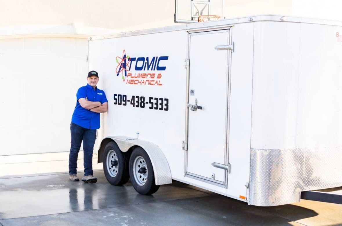 A man is standing next to a trailer that says atomic plumbing and mechanical.