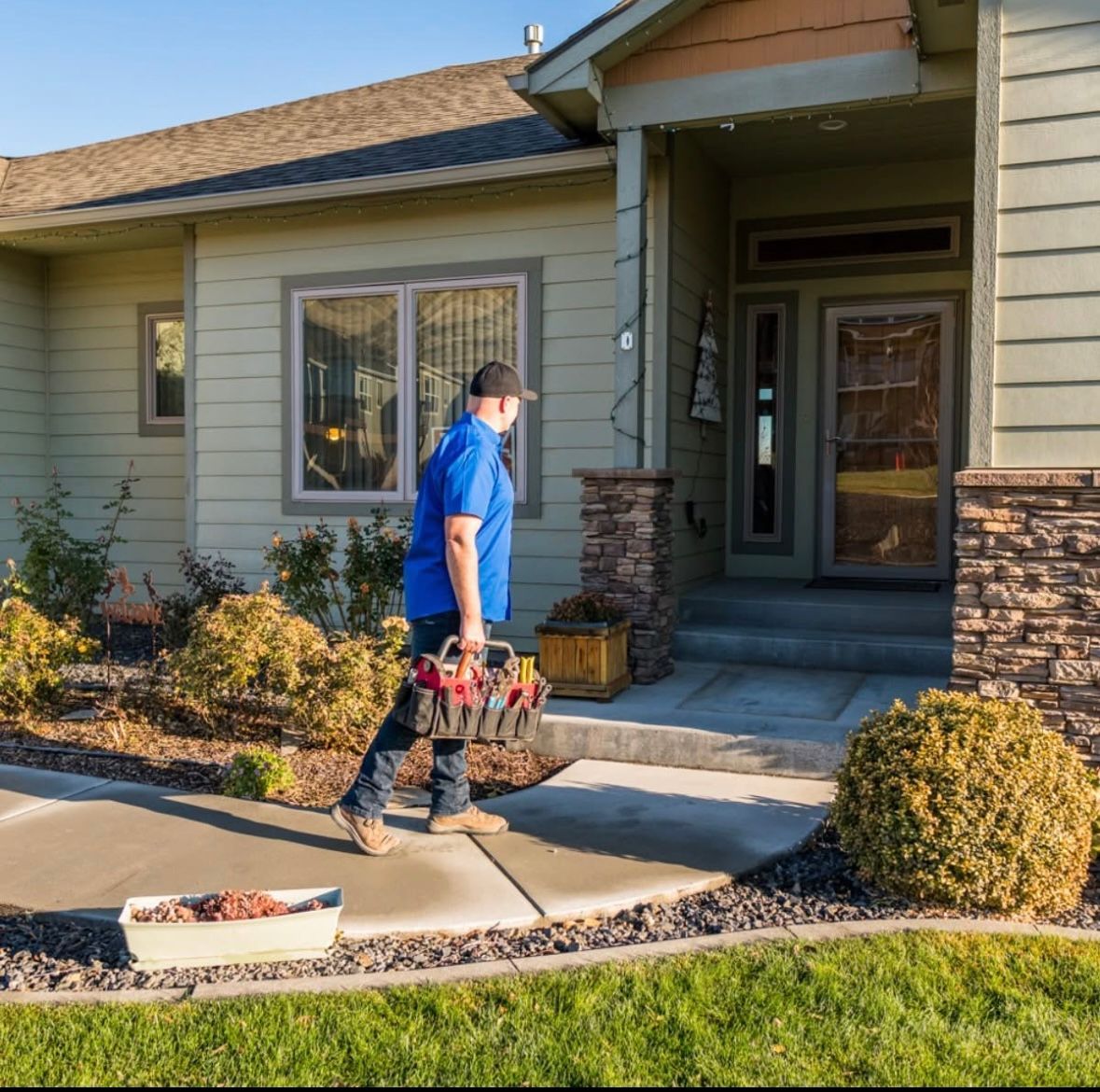 A man is walking down a sidewalk in front of a house carrying a toolbox.