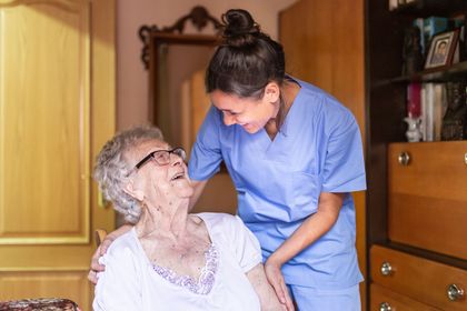 A caregiver in blue scrubs smiles at an elderly person. They are inside a home.