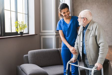 Woman in blue scrubs assists older person using a walker in a living room.