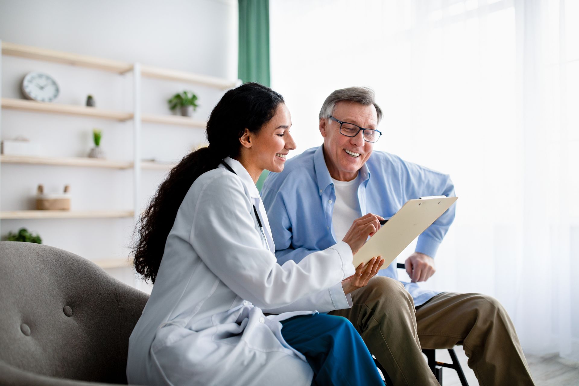 Doctor showing paperwork to a patient in a living room; smiling, holding pen and clipboard.