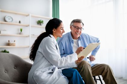 Doctor showing paperwork to a patient in a living room; smiling, holding pen and clipboard.