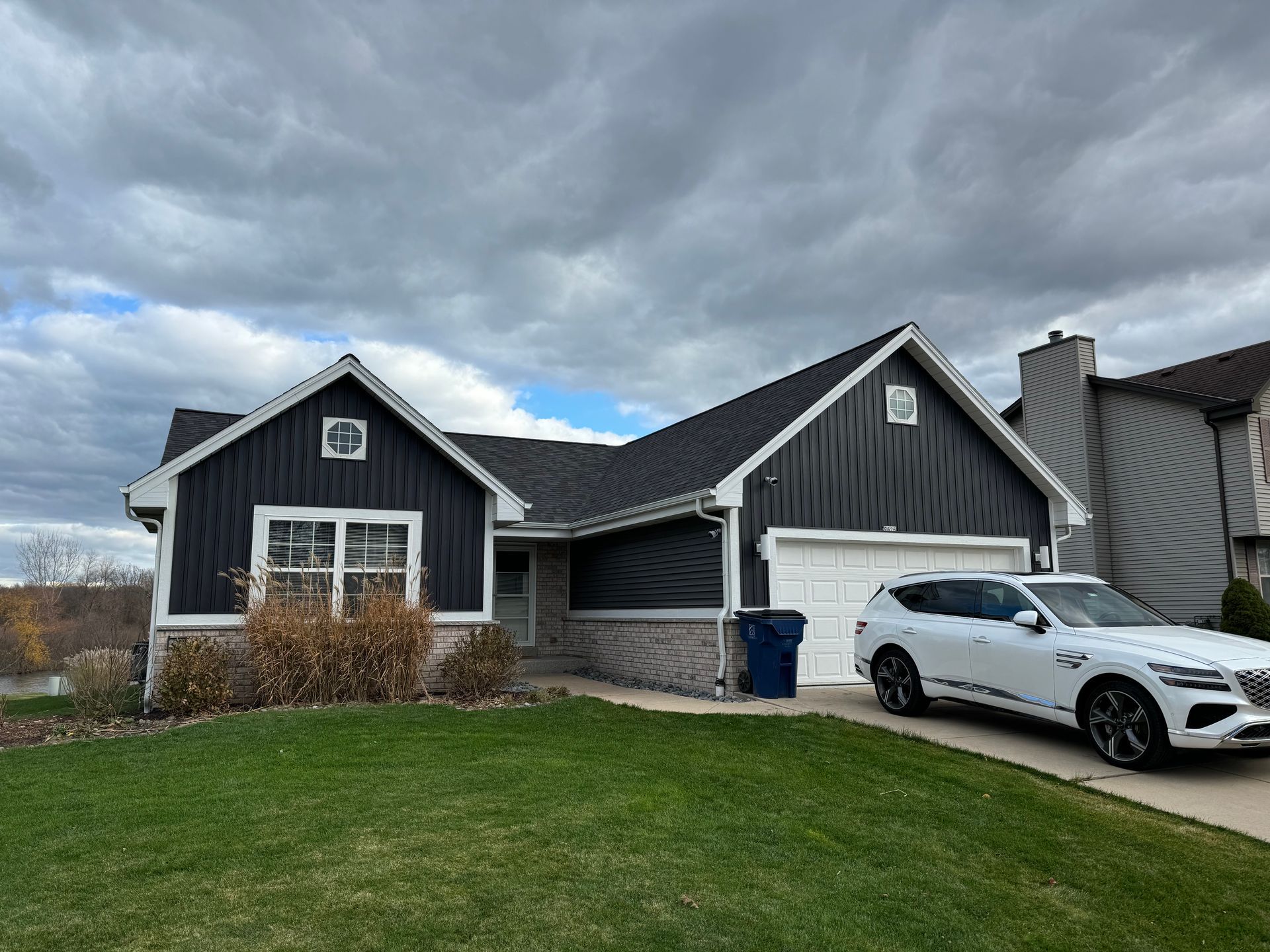 House with black siding, white garage door, and a white SUV parked in the driveway under a cloudy sky.