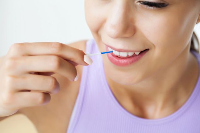 Persona che usa uno stuzzicadenti interdentale blu per pulirsi i denti, primo piano su un sorriso con una maglietta viola chiaro