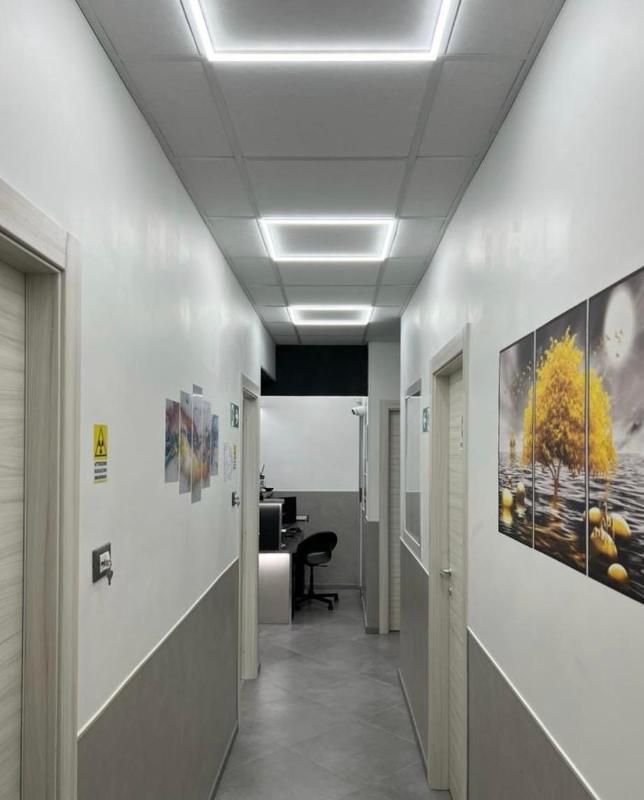 Stretto corridoio bianco con faretti a soffitto, pareti inferiori grigie e quadri incorniciati, che conduce a un'area adibita a ufficio.