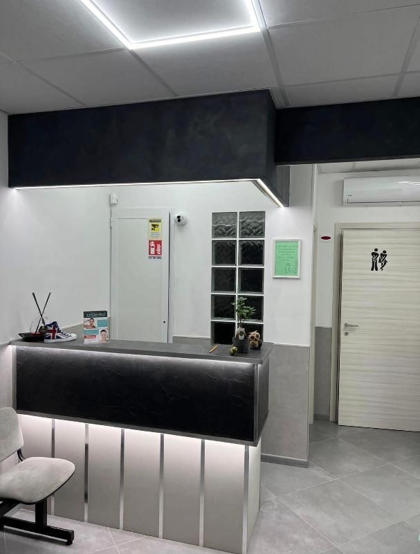 Area reception moderna con bancone nero, pareti bianche e porte dei servizi igienici sullo sfondo.