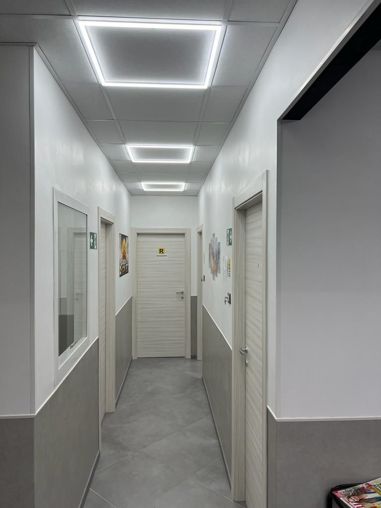 Stretto corridoio bianco con luci grigie a pavimento e a soffitto, porte su entrambi i lati che conducono a una porta chiusa.