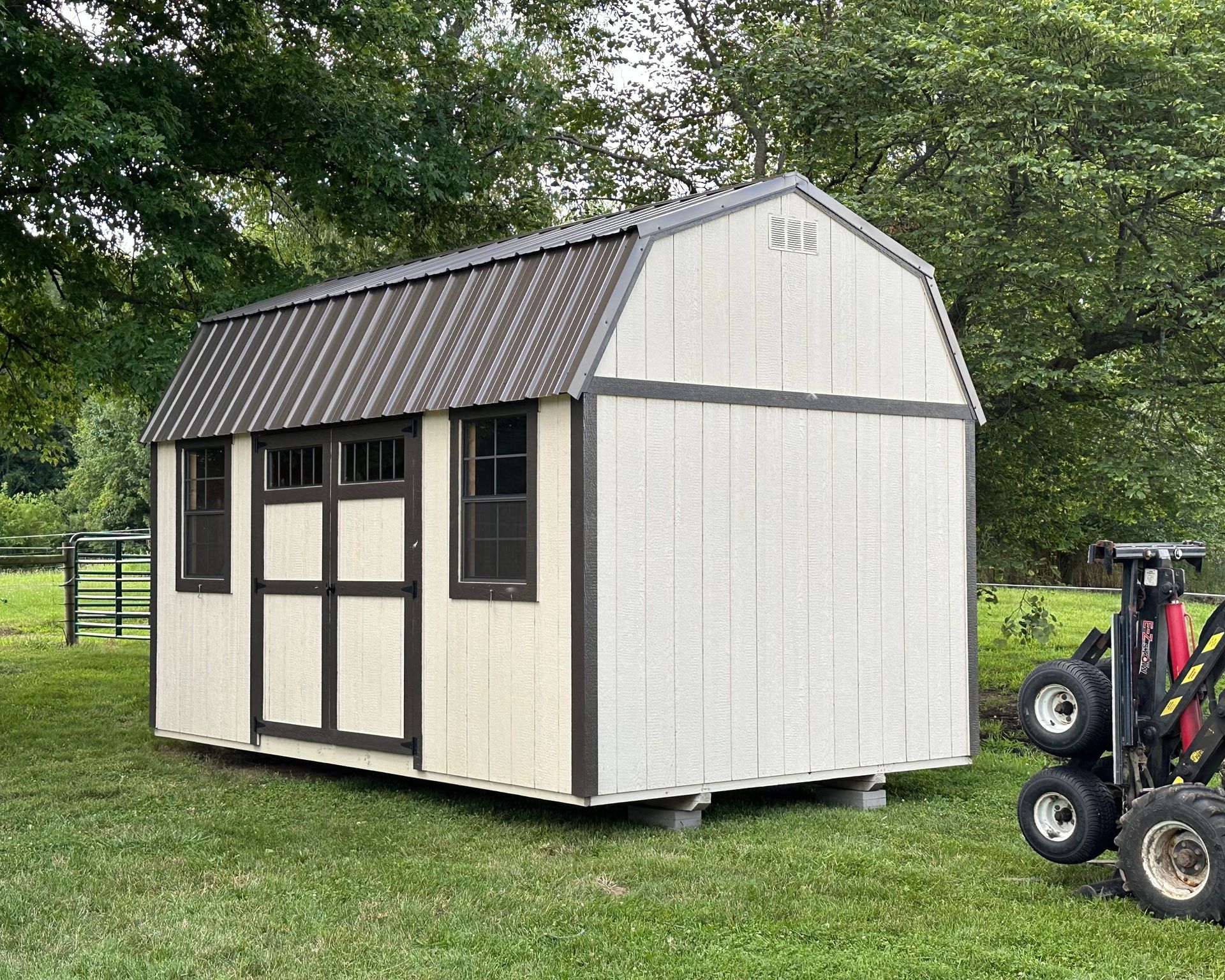 A barn shed is sitting in the middle of a grassy field next to a forklift.