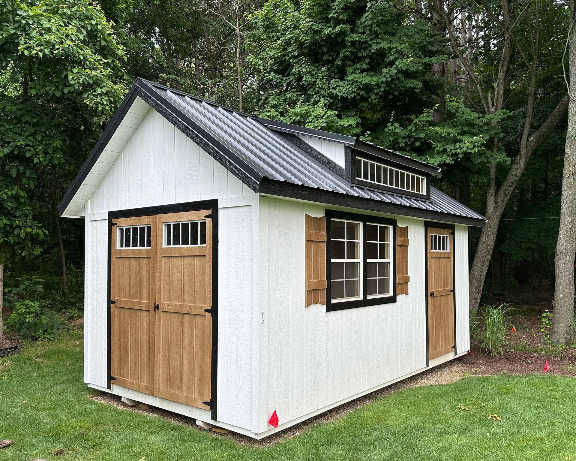 A white shed with a black roof and wooden doors is sitting in the middle of a lush green field.