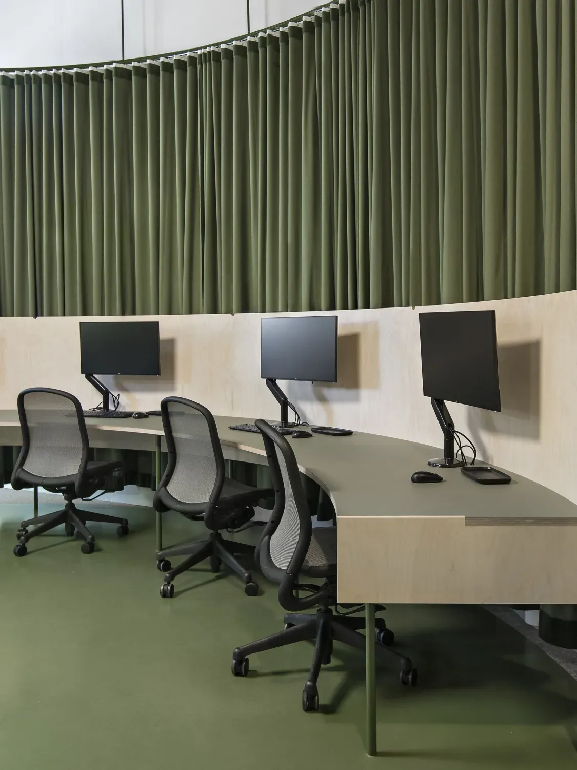 A row of desks with computer monitors and chairs in a room with green curtains.