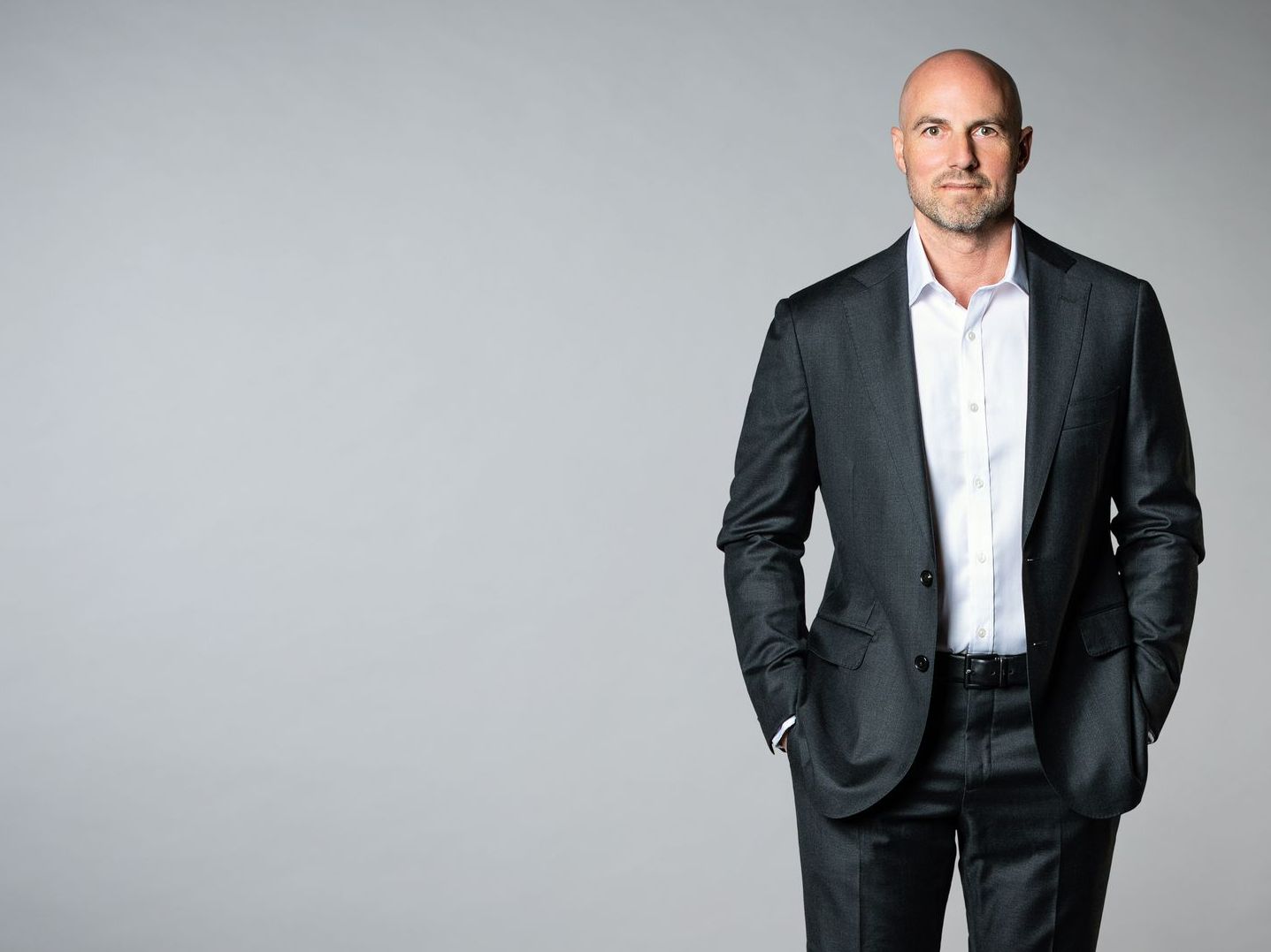 A bald man in a suit and white shirt is standing with his hands in his pockets.