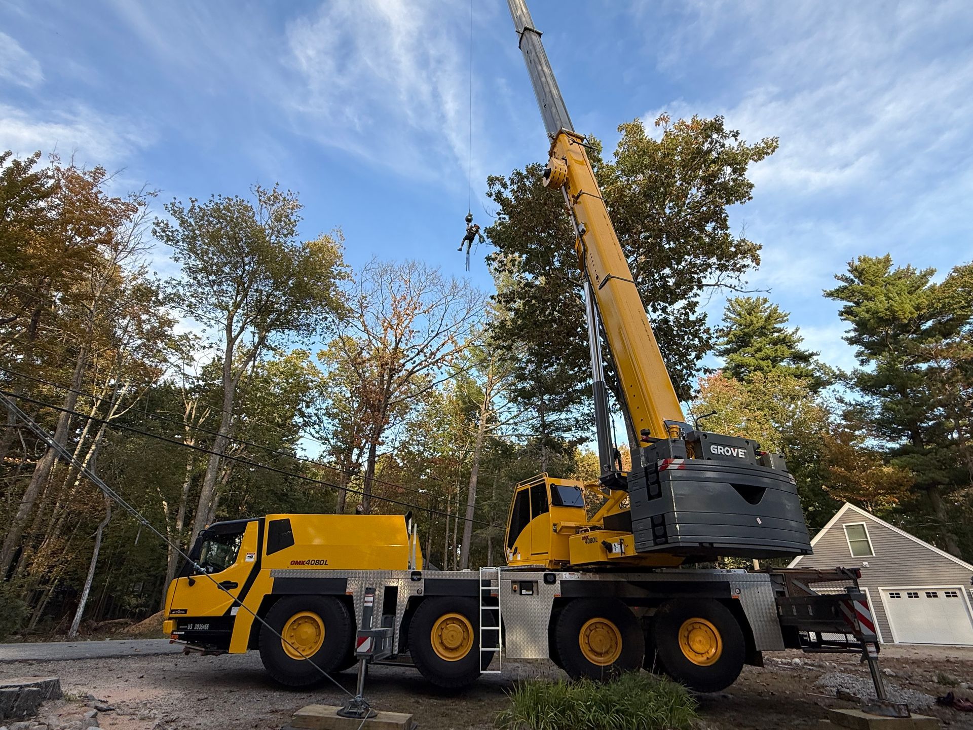 Yellow crane lifting an object outdoors, with trees and a house visible against a blue sky.