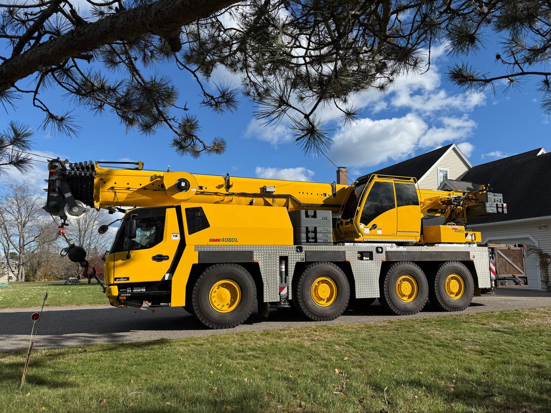 Yellow mobile crane on grass, with extended boom. Blue sky, green trees, and a house in the background.