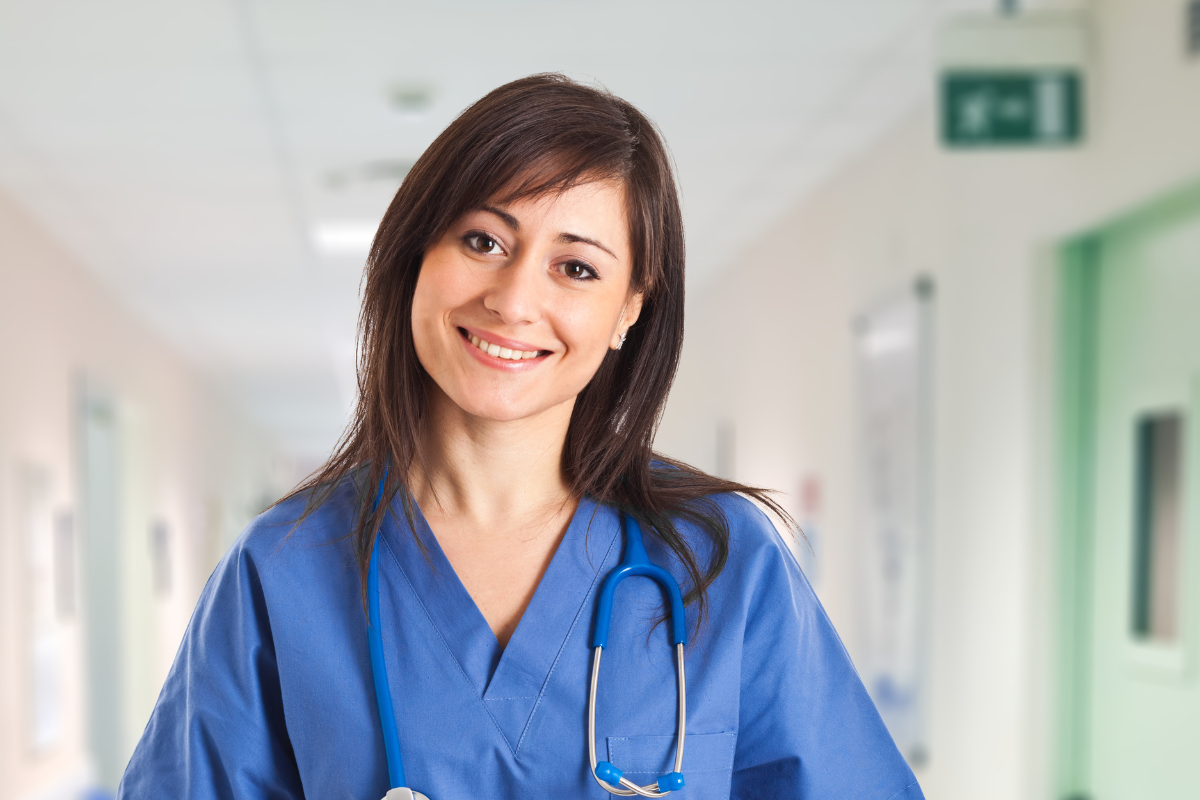 A woman in a blue scrub with a stethoscope around her neck is smiling in a hospital hallway.