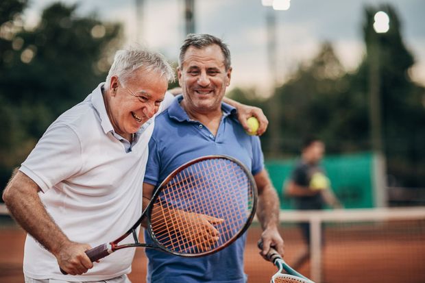 Two men laughing, holding tennis rackets on a clay court. One has a tennis ball.