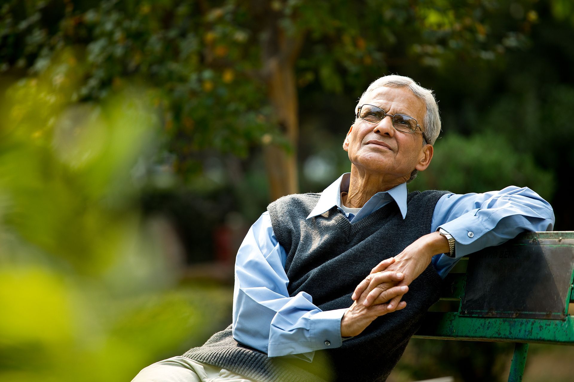 Older man wearing glasses, sitting on a park bench, looking upwards with a peaceful expression.