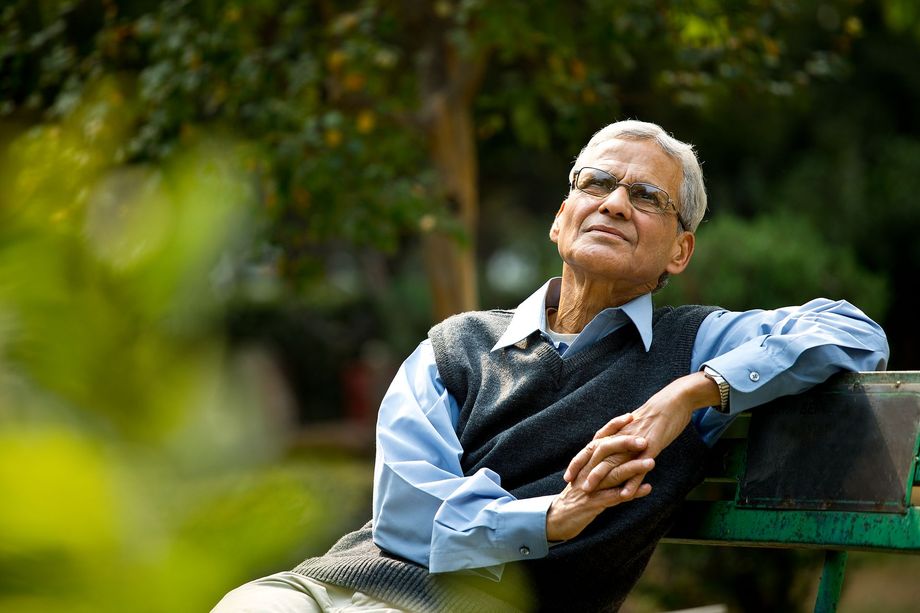 Older man wearing glasses, sitting on a park bench, looking upwards with a peaceful expression.