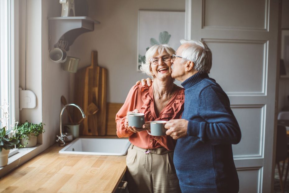 Elderly couple in kitchen, man kissing woman on cheek, holding mugs, smiling.