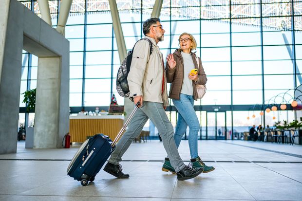 Couple walking through airport terminal, man pulling luggage, woman holding phone.