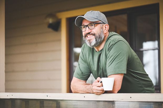 Man with glasses and a beard, wearing a cap, leans on a porch railing, holding a mug and smiling.