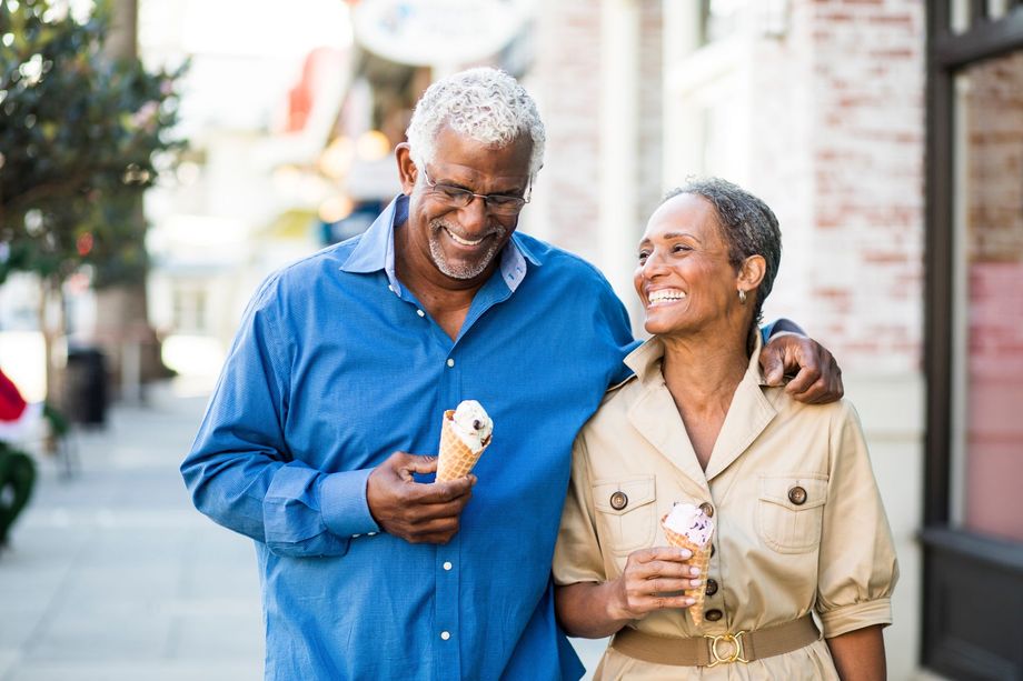 An older couple smiling, holding ice cream cones, walking down a sidewalk.