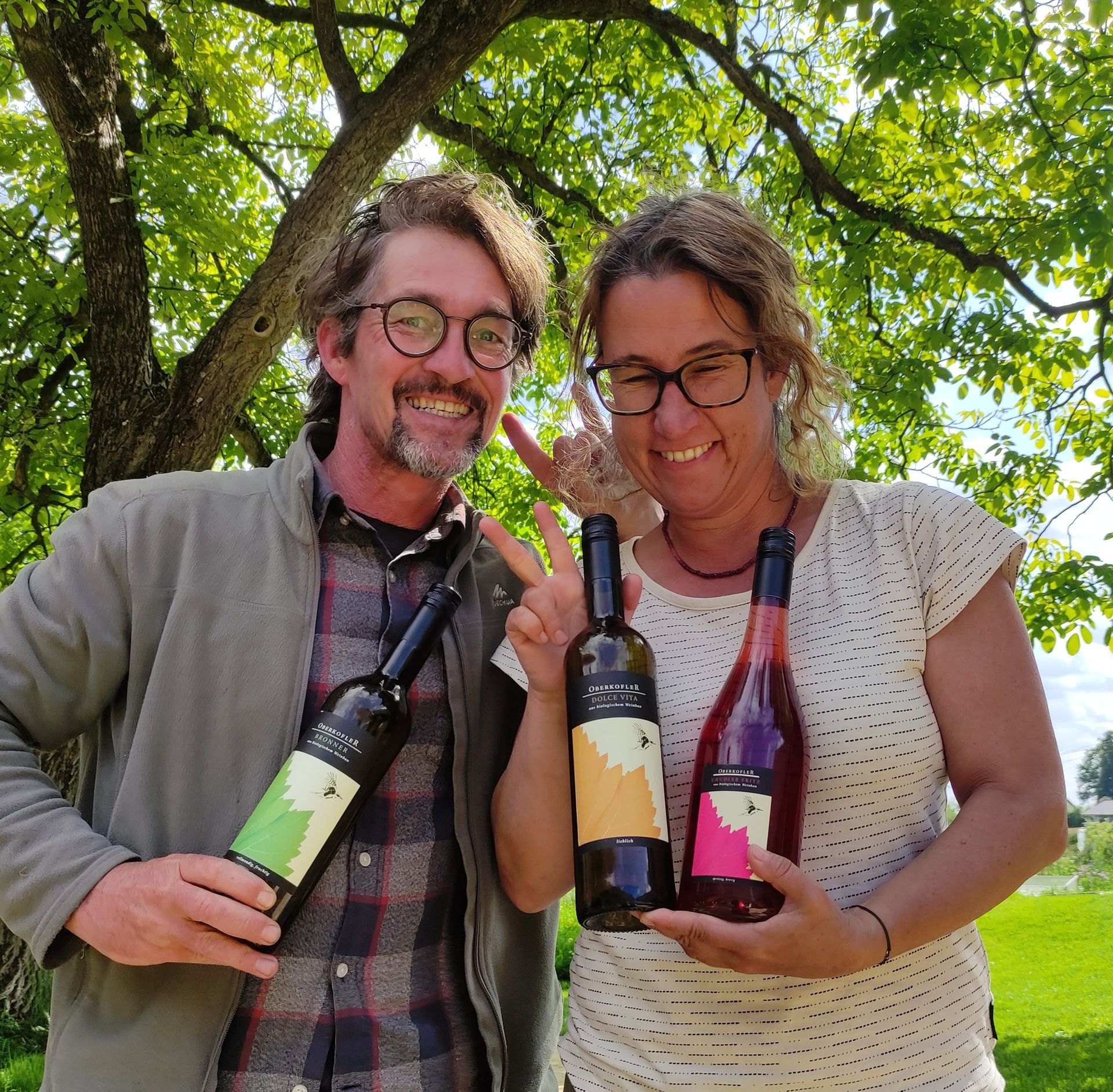 Ein lächelndes Paar hält im Freien Weinflaschen; die Frau macht ein Friedenszeichen.