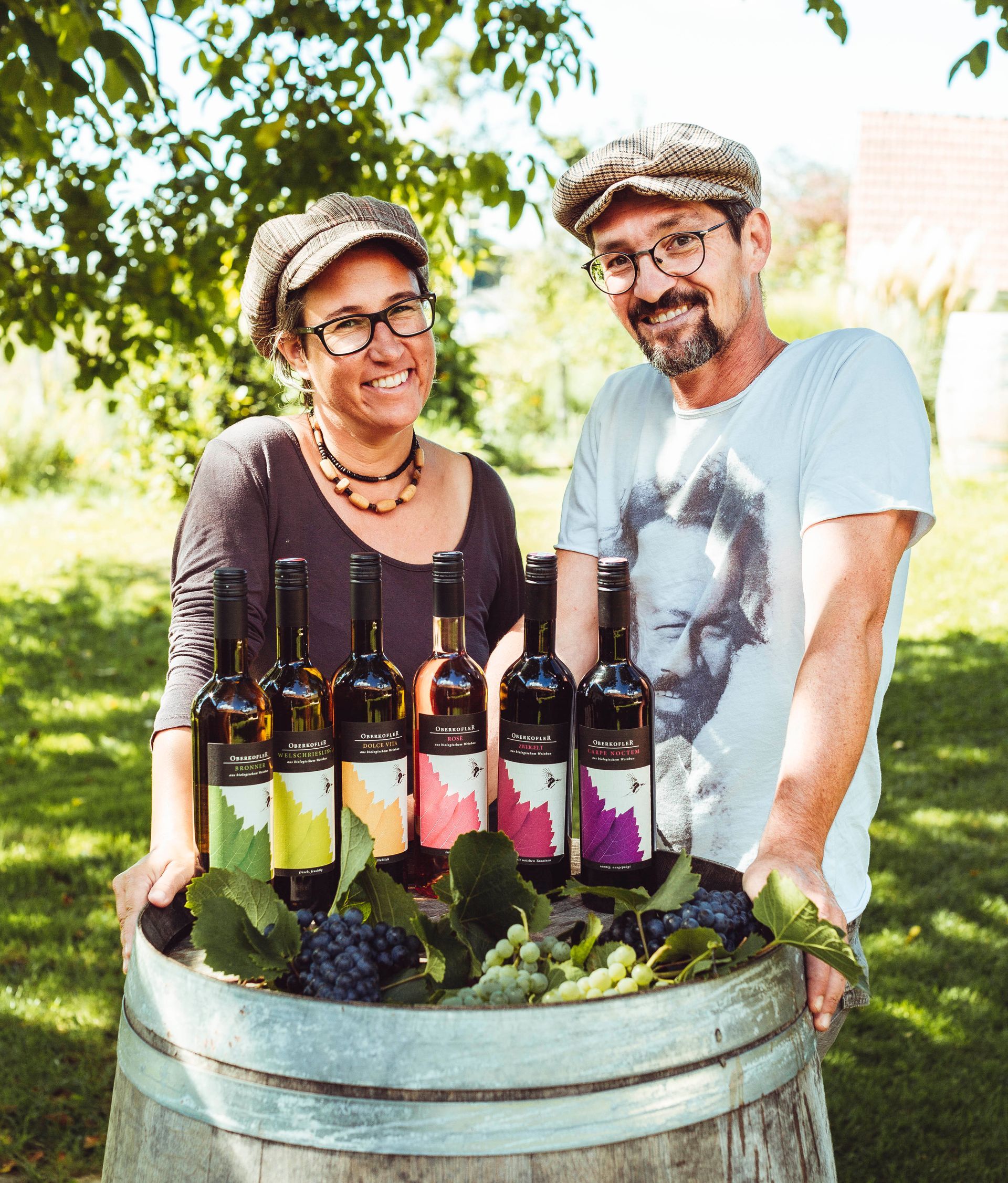 Ein Paar lächelt neben Weinflaschen auf einem Fass im Freien.