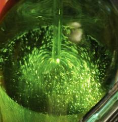 Grüne Flüssigkeit wirbelt im Inneren eines Glasbehälters, sichtbar von einer hellen Lichtquelle.