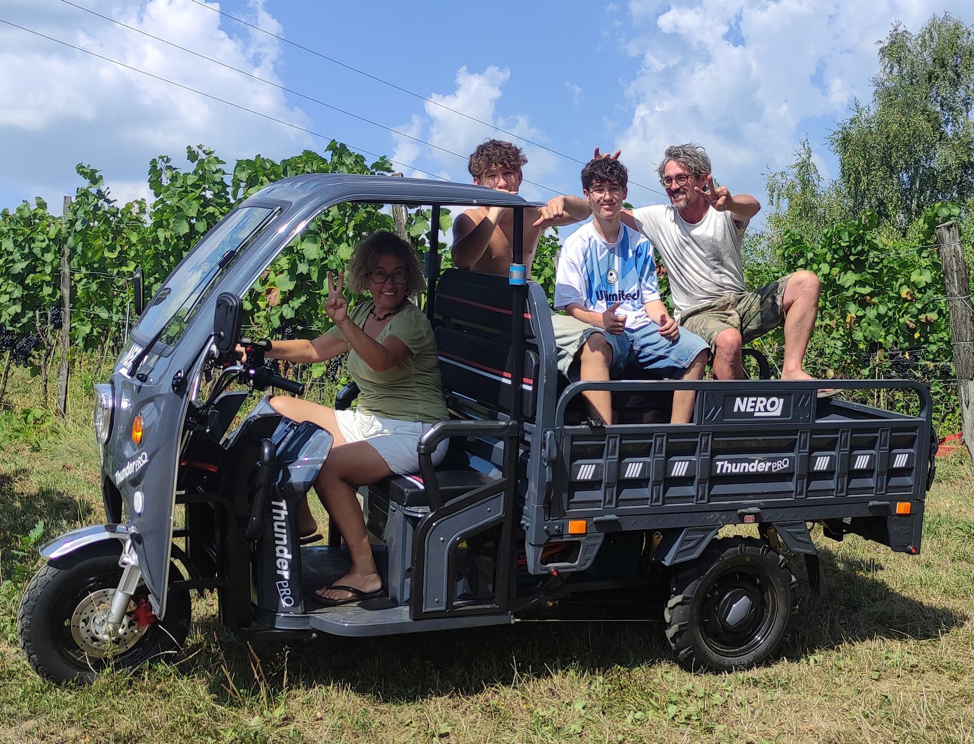 Vier Personen fahren in einem grauen, dreirädrigen Nutzfahrzeug auf einem Feld, lächeln und posieren.
