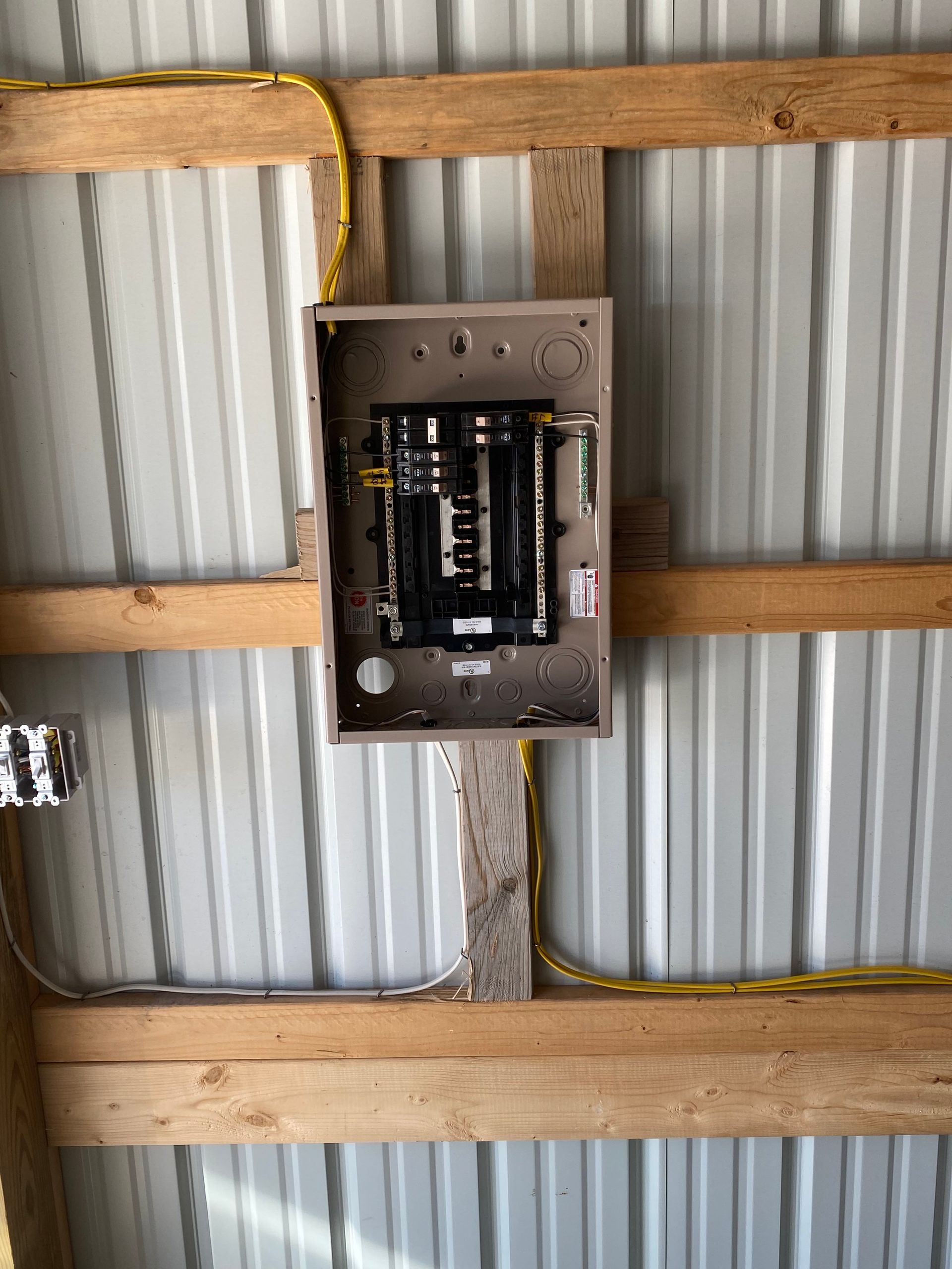 Electrical panel mounted on a wooden beam, with yellow wiring against a corrugated metal ceiling.