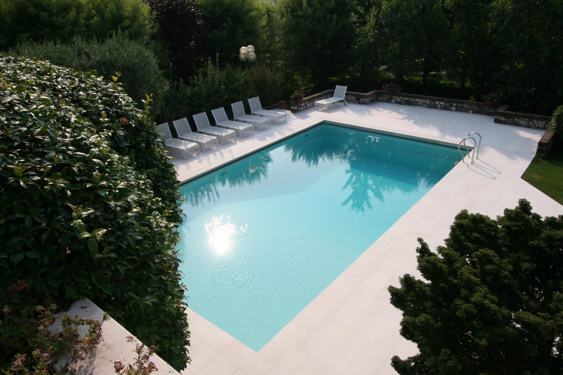Piscina circondata da un ponte bianco con sedie a sdraio e fogliame verde.