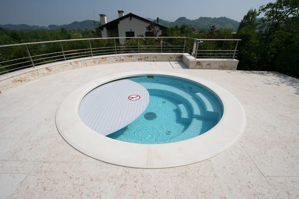Piscina esterna circolare con copertura parzialmente chiusa. Patio in pietra color crema, ringhiera in acciaio inox, casa e alberi sullo sfondo.