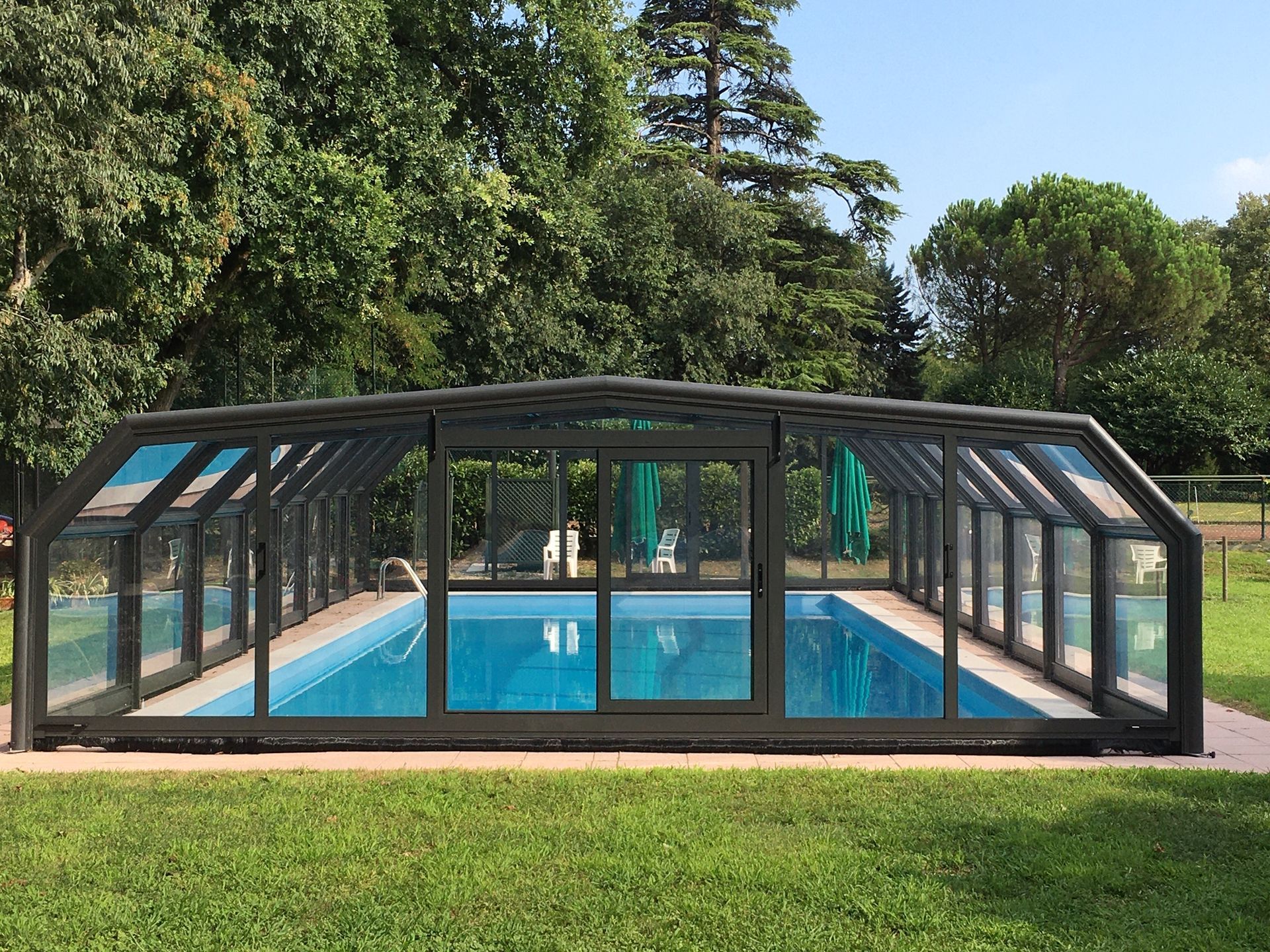 Copertura per piscina con pannelli in vetro, immersa in un prato verde, con alberi sullo sfondo.