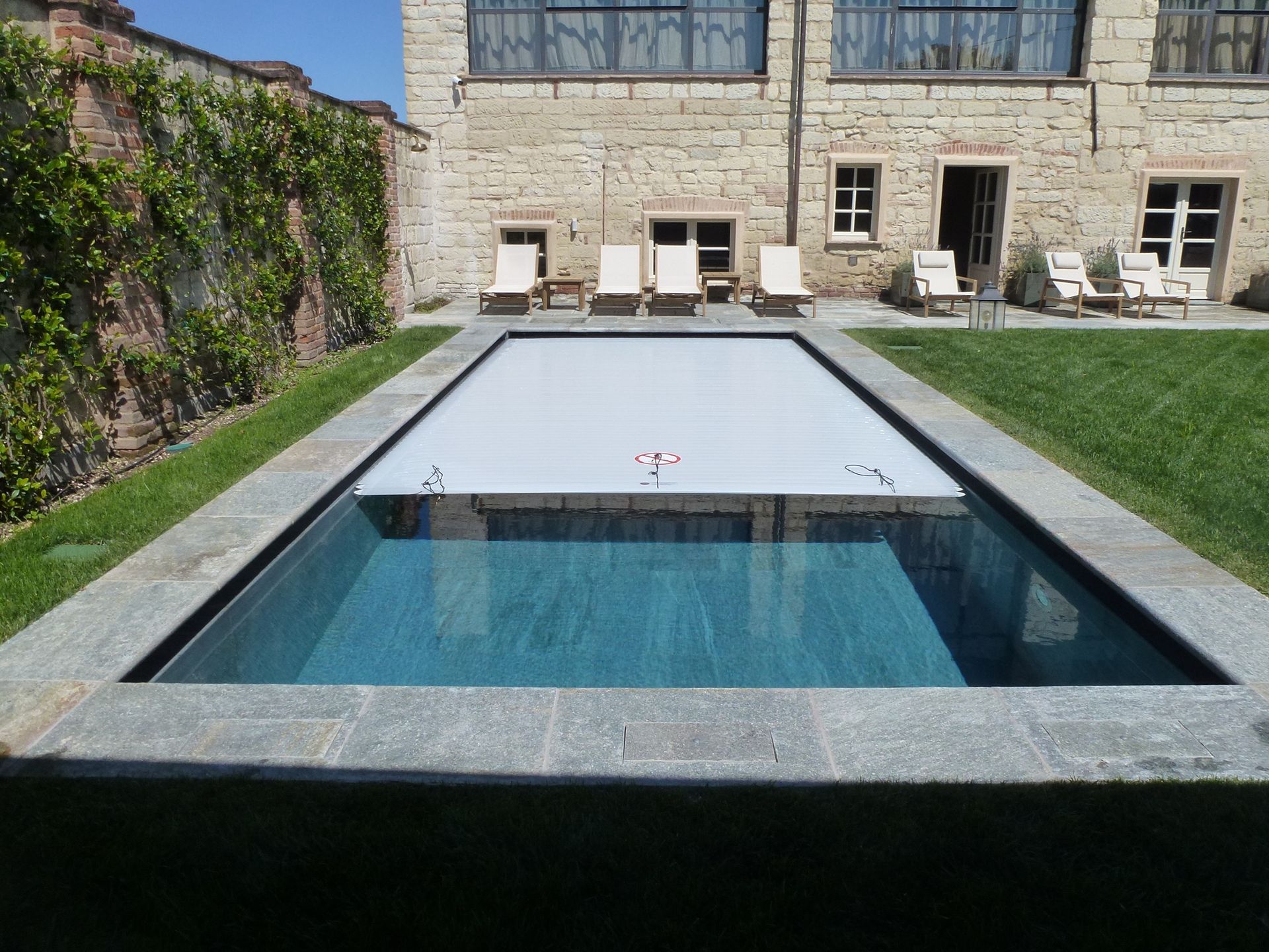 Piscina rettangolare con copertura bianca chiusa, circondata da un patio in pietra e da un prato, con uno sfondo di un edificio in pietra.