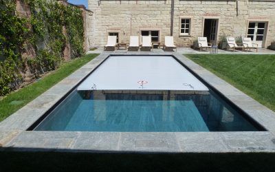 Piscina rettangolare con copertura bianca chiusa, circondata da patio in pietra, prato ed edificio.