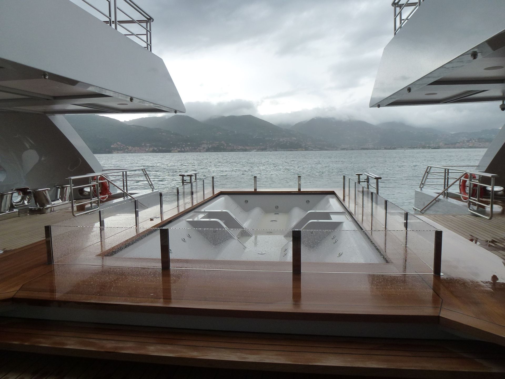 Ponte dello yacht con vasca idromassaggio, mare e montagne sotto un cielo nuvoloso.