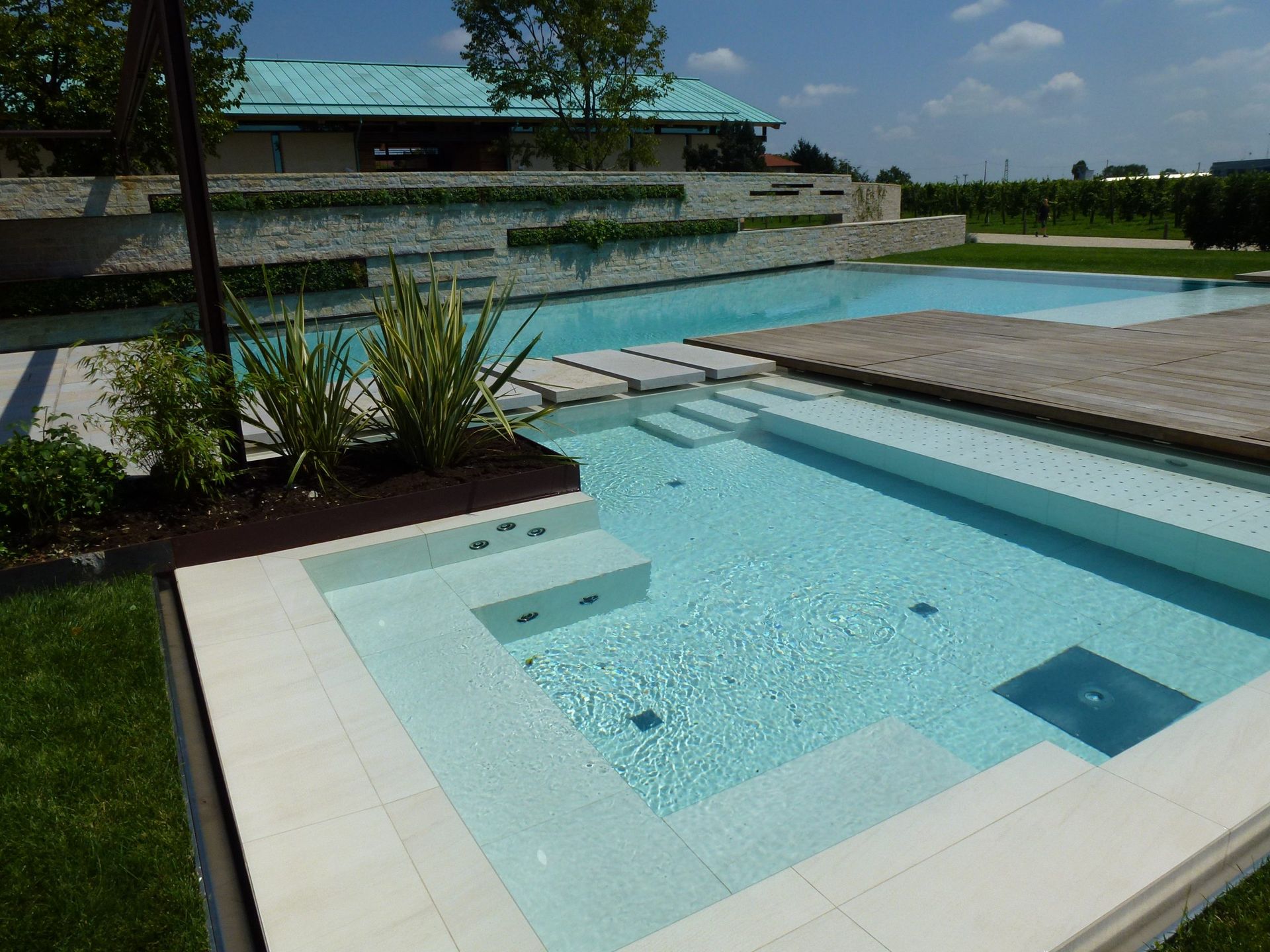 Lussuosa piscina esterna con gradini e terrazza in legno. Adiacente si trova una piscina più grande con prato verde