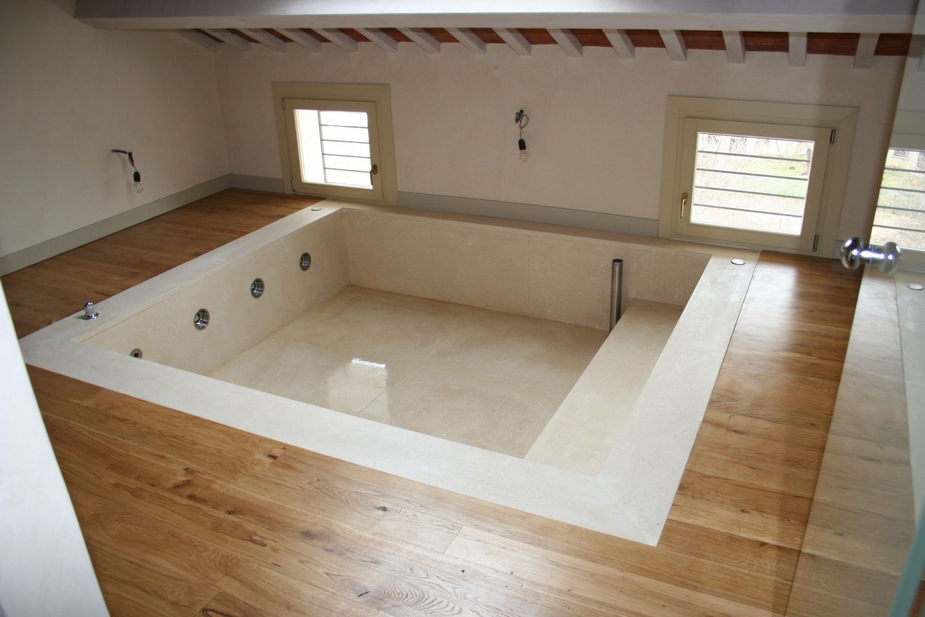 Bagno con vasca idromassaggio integrata, pareti e pavimento chiari, due finestre, pavimento in legno e travi a vista