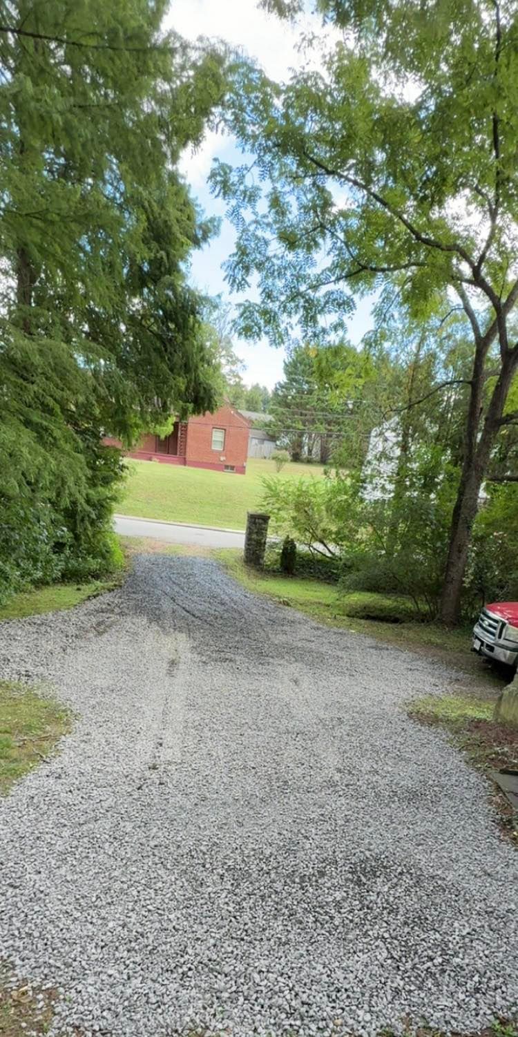 A gravel driveway leads past trees and a red building on a summer day.
