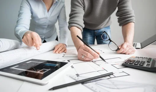 Two people reviewing floor plans and tablet data at a desk with a calculator and glasses