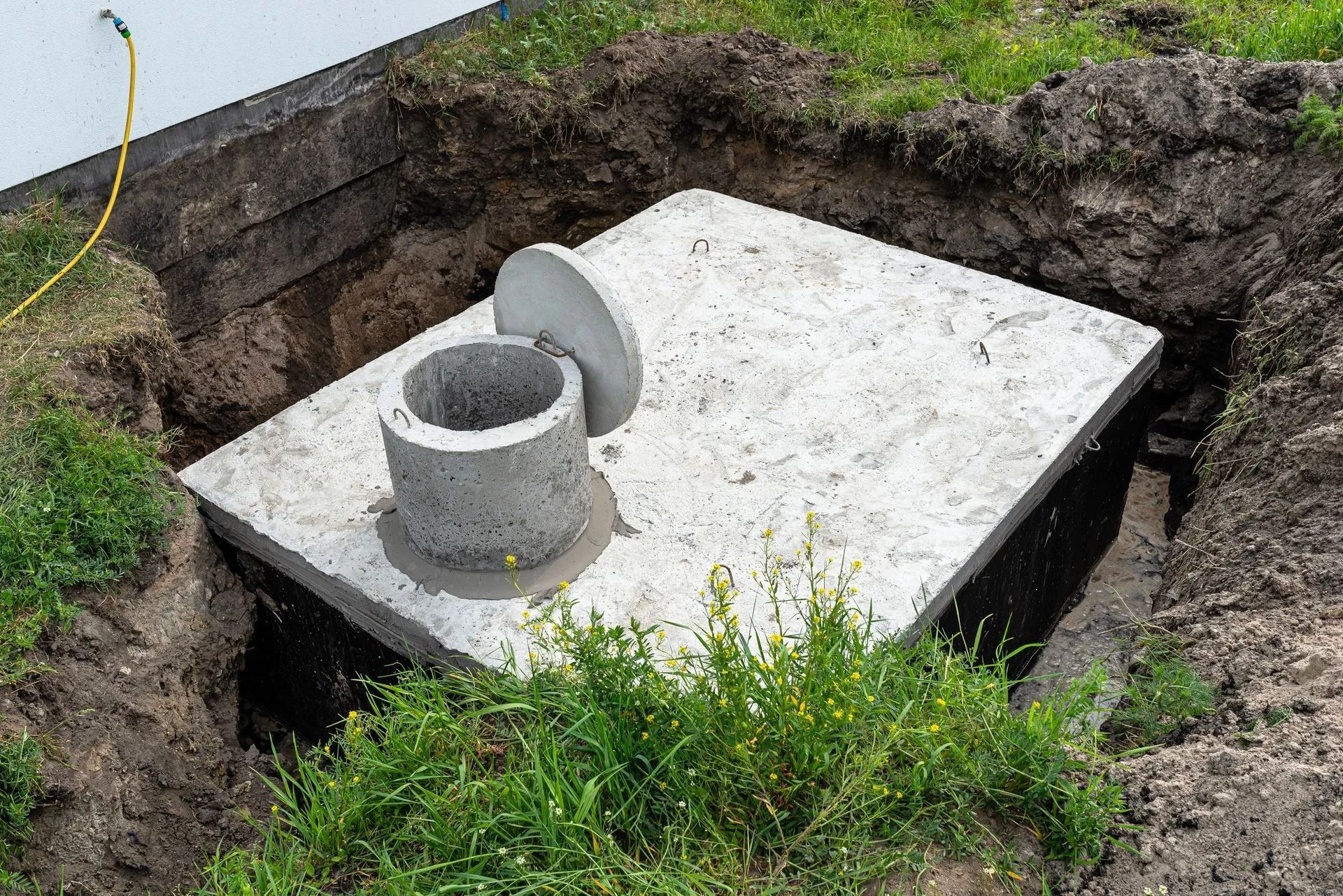 A square concrete septic tank cover with an open circular access port set into a dug-out hole in the ground.