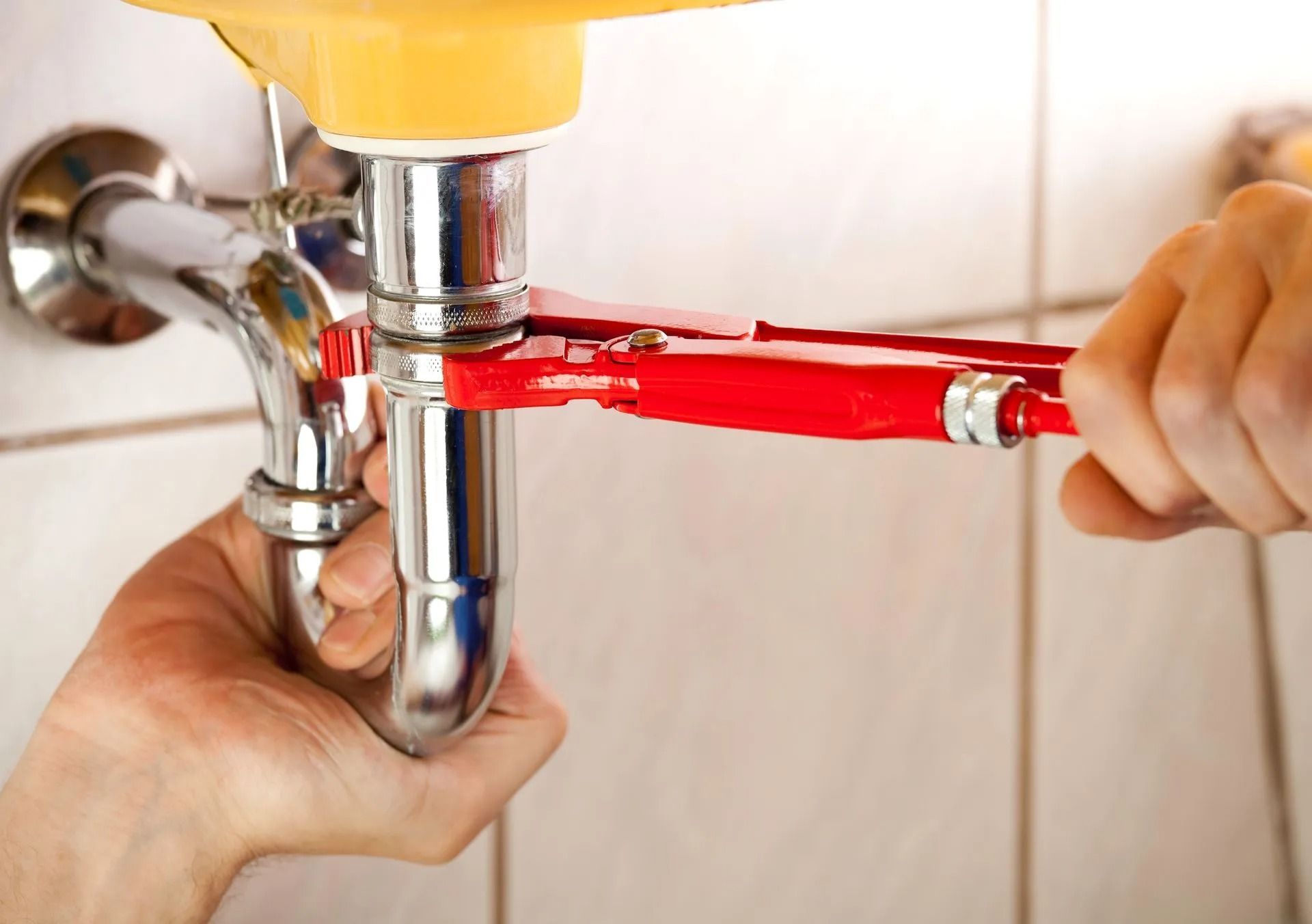 A person uses a red wrench to tighten a chrome p-trap pipe under a sink.