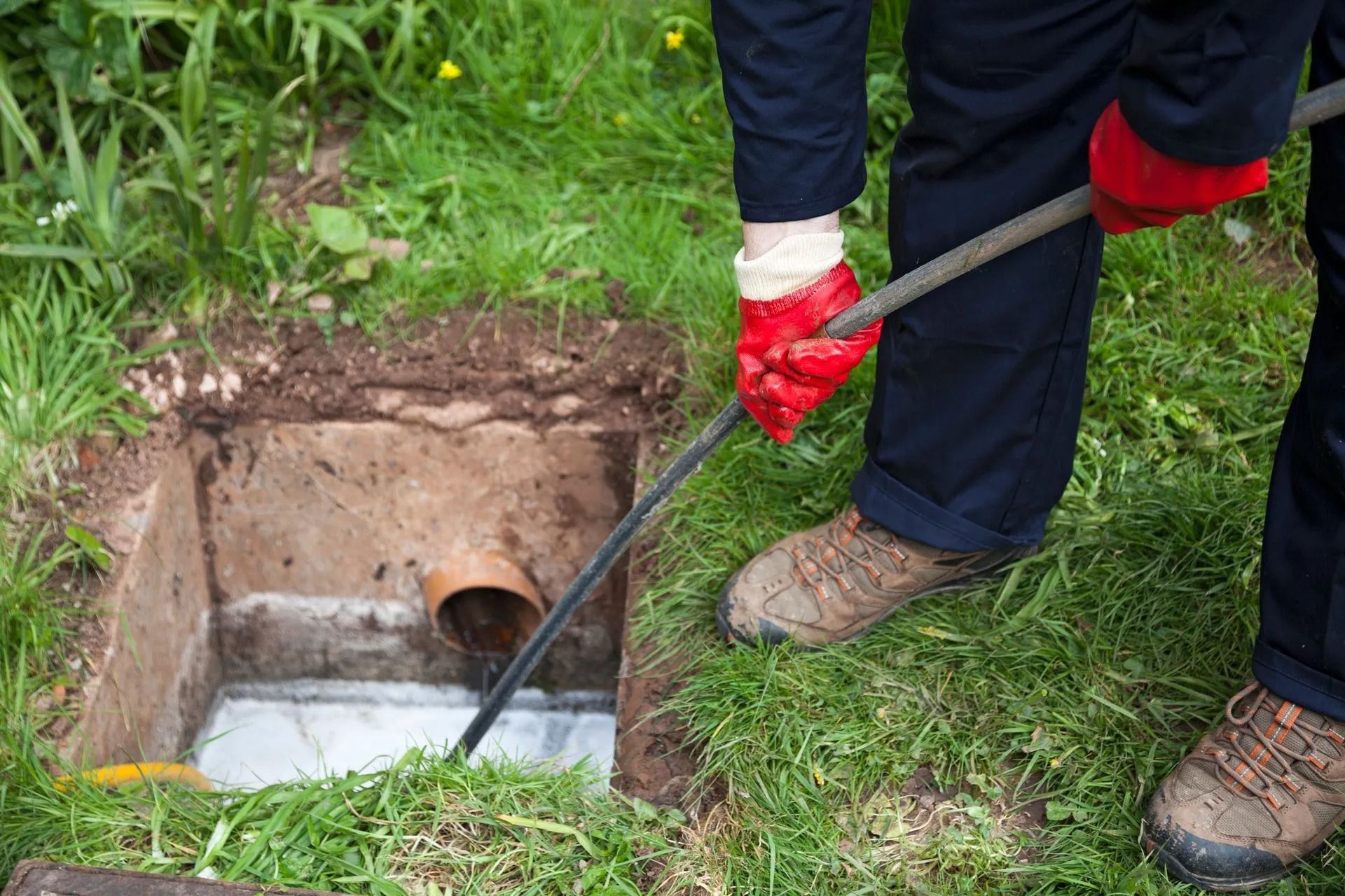 A person wearing work clothes and red gloves uses a plumbing snake to clear a clog in an outdoor drainage pit.