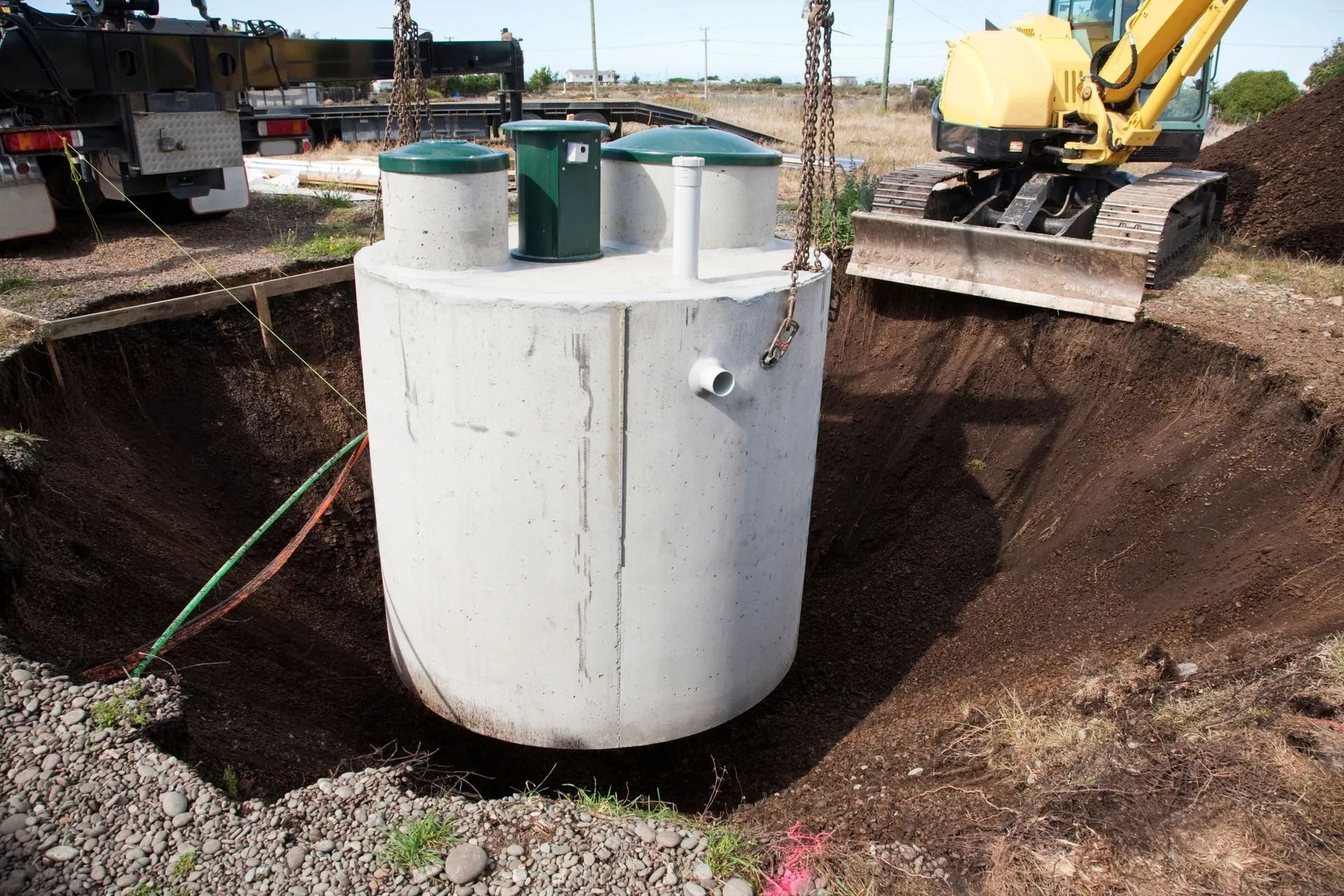 A concrete septic tank is being lowered into a excavated circular pit by a construction crane on a sunny day.