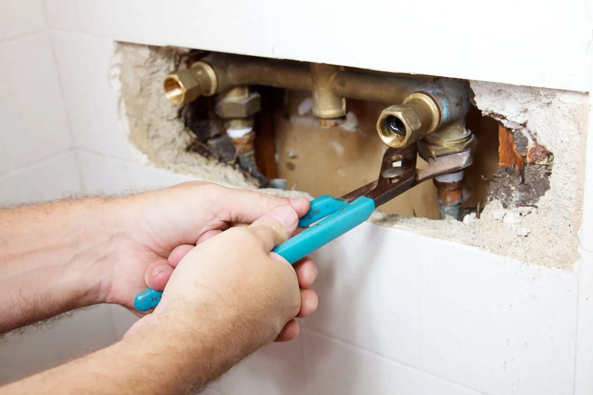 A person uses teal-handled pliers to tighten a brass pipe fitting inside an opening in a tiled bathroom wall.