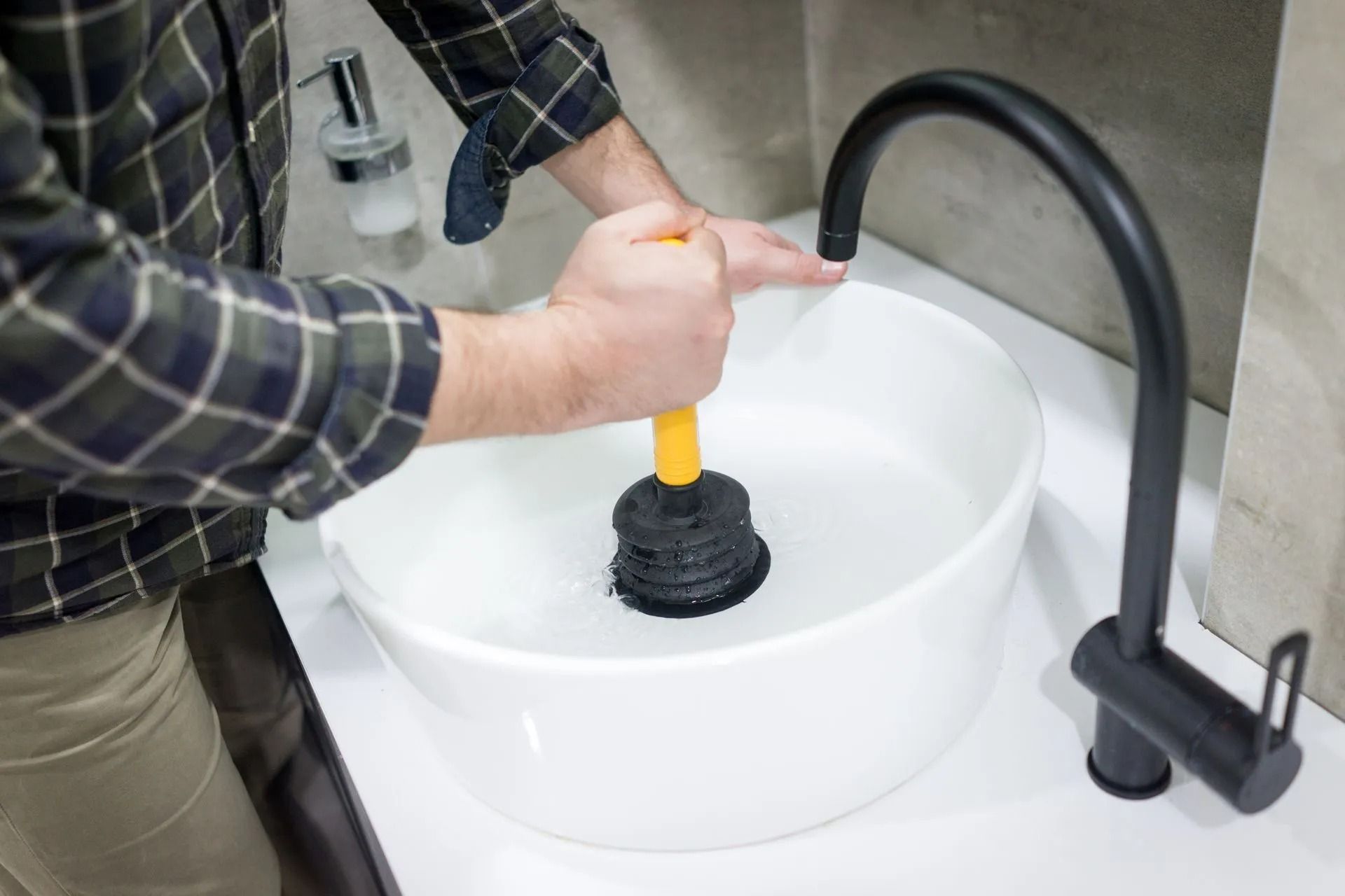 A person in a plaid shirt uses a yellow-handled black plunger to clear a drain in a white bathroom sink.