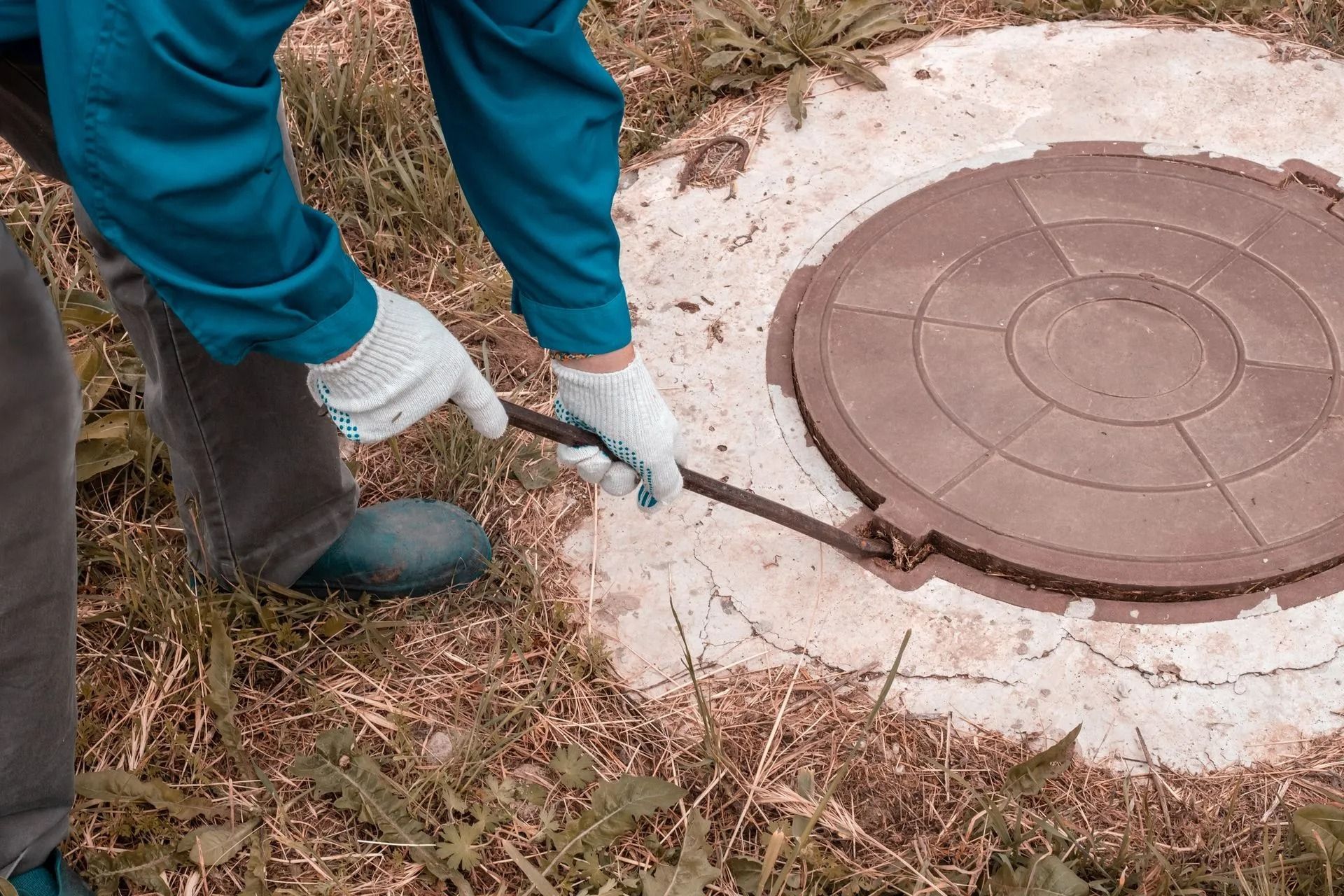A person wearing gloves uses a pry bar to lift a heavy, circular brown manhole cover set in concrete on the ground.