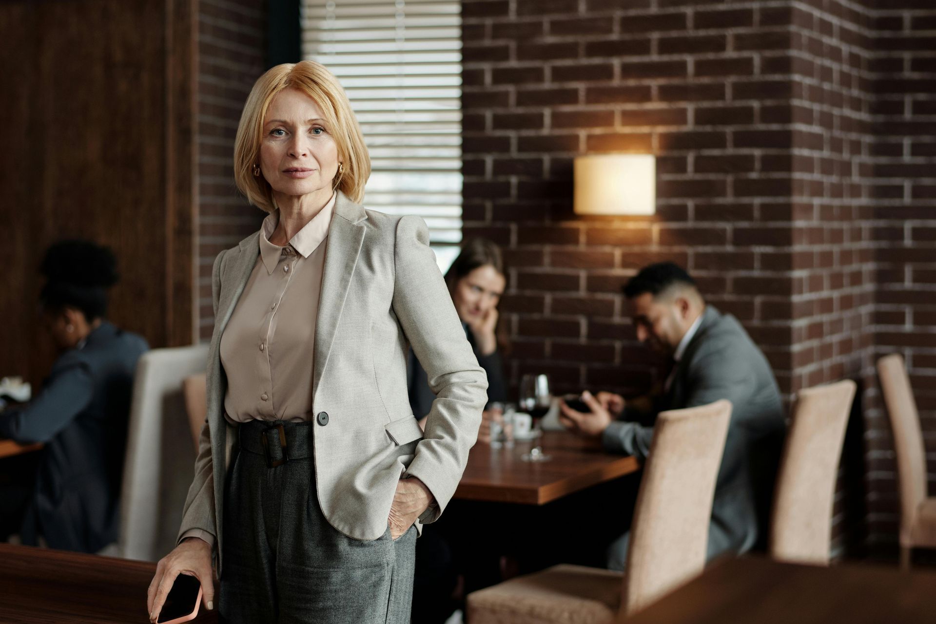 Woman in blazer standing in a restaurant; other patrons in the background.